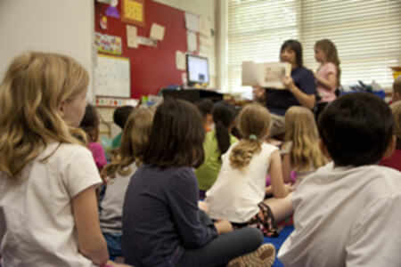 Bilde av barn som sitter på gulvet i et klasserom mens læreren i front holder opp en bok.