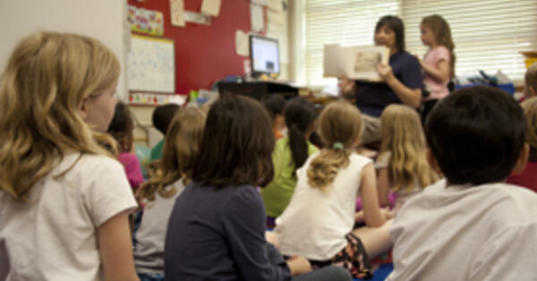 Bilde av barn som sitter på gulvet i et klasserom mens læreren i front holder opp en bok.